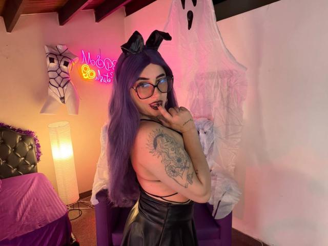 LilithDark69 with purple hair, glasses, black bunny ears, and a tattoo on her arm, touching her lips, topless, showing her tits, in a pink-lit room with a bed.