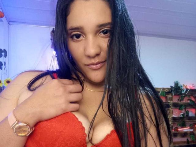 CristalRios with long black hair, red lace bra, showing her tits, slight smile, pink wristwatch, gold necklace, in a room with blue lighting.