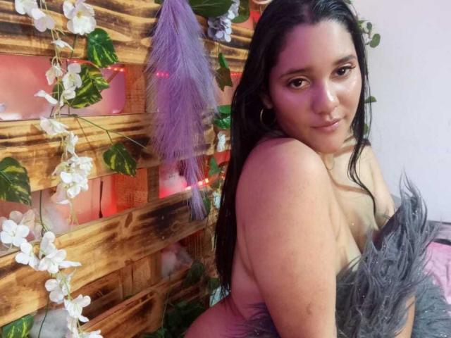 CristalRios, senza top, con lunghi capelli neri, occhi marroni e un leggero sorriso, tiene in mano della pelliccia grigia, mostrando il seno, davanti a un muro di legno con fiori bianchi e piume viola..