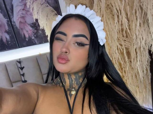 LairaHearvy with black hair, white maid headband, thick black eyebrows, tattooed neck, and pouty lips, winking, showing off her boobs. She's wearing a black choker with a gold ring.
