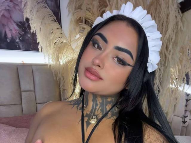 LairaHearvy with long black hair, white maid headpiece, thick black eyebrows, eyeliner, pink lipstick, tattoos on her neck, and a nose ring, looking at the camera.