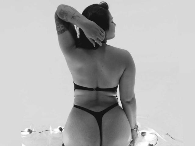 Black-and-white photo of BonnieGray, back to camera, wearing a thong and bra, showcasing her round ass, tattoos on right arm, and short dark hair, with a string of lights.