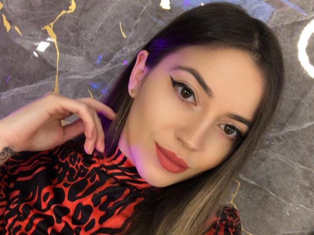 Lesysl with long brown hair, thick eyebrows, and red lipstick, wearing a leopard print red and black top, showing off a tattoo on her right wrist.