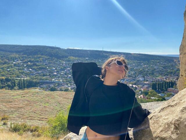 LeScharwath, com cabelo castanho curto, óculos de sol, cropped preto e jaqueta preta, em pé em uma rocha, céu azul, reflexo do sol, vestindo jeans azuis, olhando para cima..