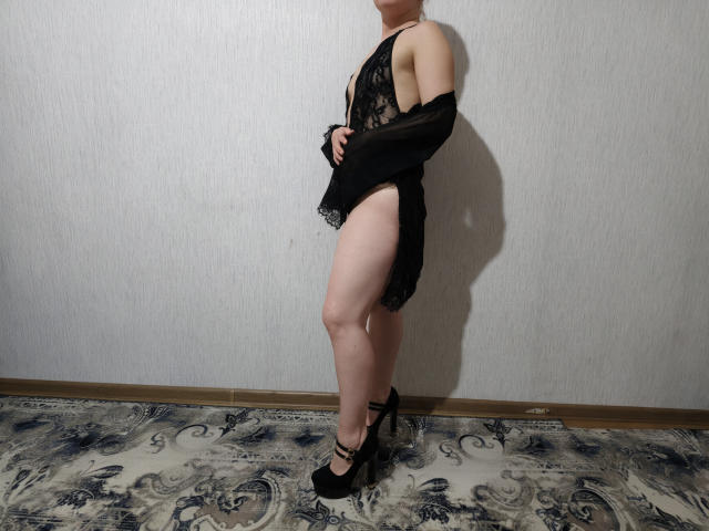KaterinCold in black lace lingerie, exposing her tits and ass, with a black shrug, standing in black high heels, pale skin, and a short hairstyle, on a patterned rug.