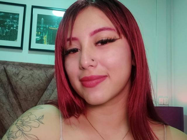 MaddyMills a les cheveux roux, des cils épais, des sourcils roses, un anneau au nez et un tatouage floral sur son épaule. Elle sourit, avec du rouge à lèvres rose, et porte un haut à fines bretelles..