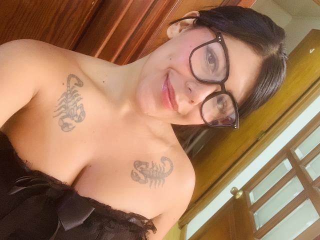 CatalinaReiyna with black glasses, smiling, has tattoos on both her tits, wearing a black lace off-shoulder top, in a selfie. She has medium skin and black hair.