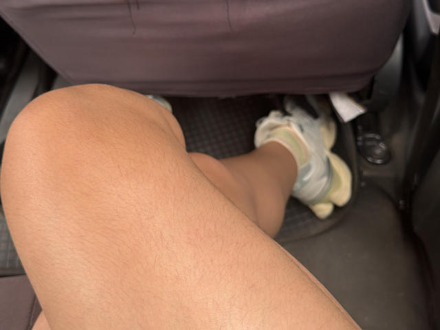 MiiaPalmer's tan, hairy, bent leg with a white sneaker and green sock under a car seat. Visible foot and shoe. Textured black car floor.