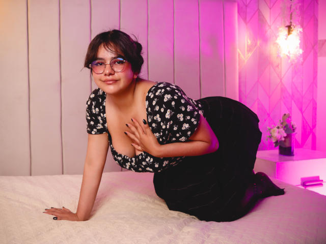 Nicole Santana, usando óculos, com uma blusa preta estampada com flores e mangas bufantes, está de quatro em uma cama branca, com a mão sobre os seios, usando uma saia preta e com um sorriso sedutor..