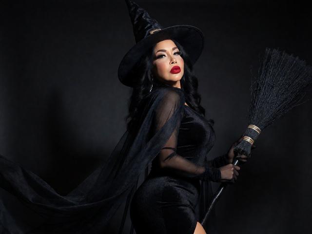 AngelinaDeleon with black witch hat, sheer gloves, and dress, holding a broom, has red lips, wavy black hair, and is showing off her big tits and ass.