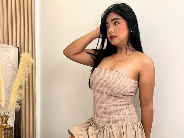 VictoriaKross, con cabello negro largo, vestida con un vestido beige sin tirantes, con la mano en el cabello, mirando hacia un lado, labial rojo, maquillaje sutil, un collar pequeño, piel morena, senos pequeños, linda..