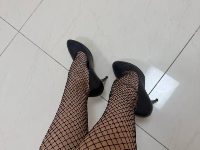 Le gambe di MoonLiza, indossanti calze a rete nere e tacchi alti neri, sono in piedi su un pavimento di piastrelle bianche. I suoi tacchi sono puntati verso l'alto, mostrando le suole..