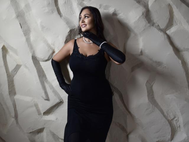 MilaUrban with dark hair, wearing a black lace dress, black gloves, and a necklace, standing against a textured wall, hand near her chin, looking to the side, tits visible.
