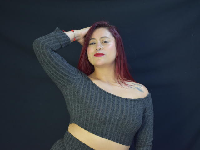 Evans Samantha, com cabelo ruivo, vestindo uma cropped cinza texturizada, expondo seus seios e uma tatuagem no ombro direito, com a mão no cabelo, fundo preto, pulseira vermelha..