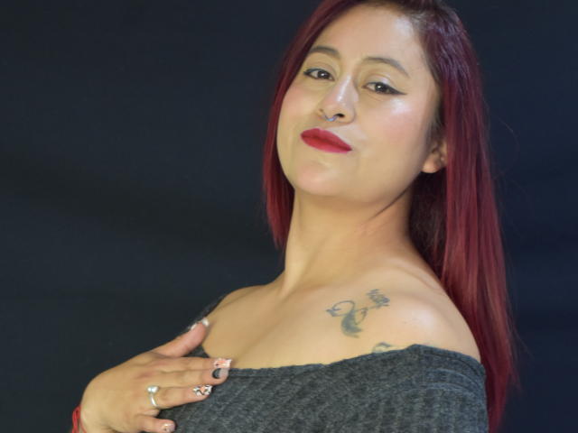 EvansSamantha with red hair, nose ring, red lipstick, grey off-shoulder top, tattoos on shoulder, holding chest, ring on finger, black background. Boobs, tits, hand, nails, ring.