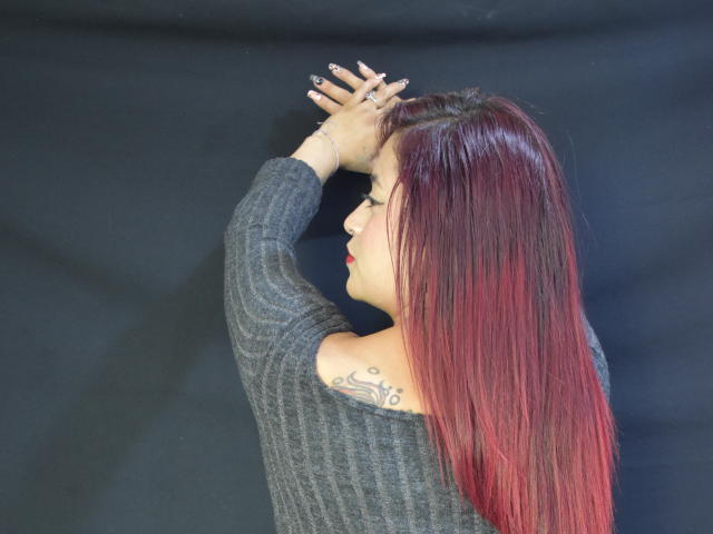 Evans Samantha, com cabelo ombré de vermelho para preto, vestindo um suéter cinza de malha canelada, braços levantados, mostrando uma tatuagem na omoplate esquerda, e batom vermelho..