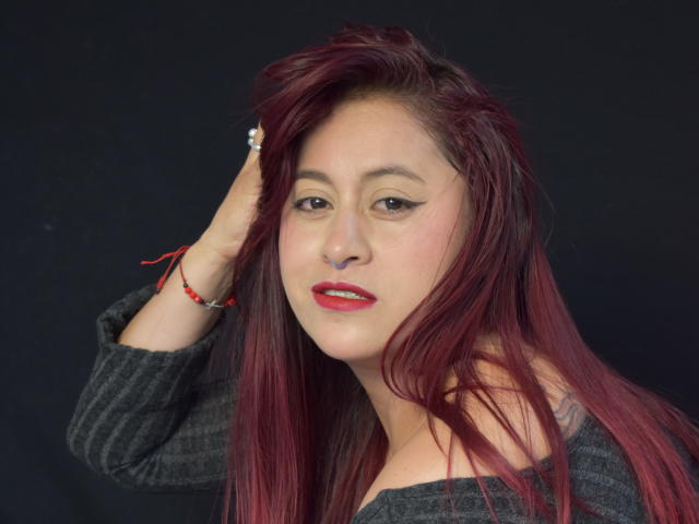 EvansSamantha, con lápiz labial rojo, maquillaje de ojos oscuro y cabello largo rojo, vestida con un suéter gris de corte asimétrico, con la mano en el cabello, fondo oscuro, pulsera roja y cuenta blanca..