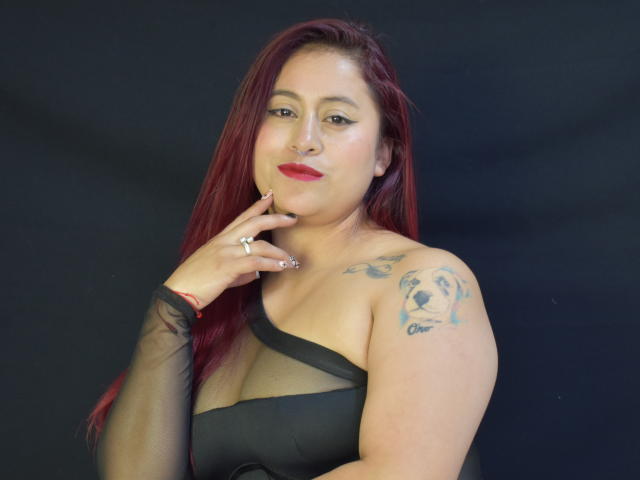 EvansSamantha, con cabello rojo, vestido negro asimétrico, tatuajes en el hombro, lápiz labial rojo, anillo en el dedo, fondo negro, tocándose la barbilla, mirando a la cámara, sonriendo ligeramente..