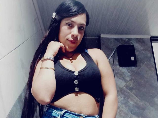 ValentinaRicci tem cabelos pretos longos, uma pequena flor branca no cabelo, veste uma cropped preta com detalhes de botões, mostrando seus seios, e jeans azuis em uma selfie..
