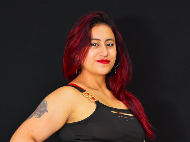 Evans Samantha, con cabello rojo, lápiz labial rojo, camiseta sin mangas negra y un tatuaje en su brazo derecho, sonriendo a la cámara, mostrando sus pechos de tamaño medio..