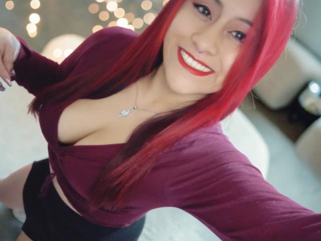 Evans Samantha, met rood haar, lachend, gekleed in een diep bordeauxrode top, waarbij haar grote borsten zichtbaar zijn, zwarte shorts en rode lipstick, die een selfie maakt, met bokeh-lichtjes op de achtergrond..