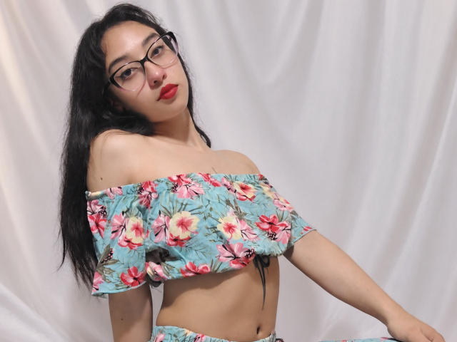 CamiVargas, con lunghi capelli neri, occhiali e rossetto rosso, indossa una blusa floreale con una spalla scoperta, che mette in mostra il suo seno piccolo e un laccetto nero, su uno sfondo bianco..