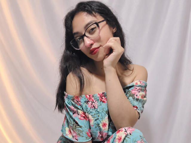CamiVargas con gli occhiali, capelli neri ondulati, rossetto rosso, vestito floreale con una spalla scoperta, mano sul viso, sguardo leggermente di lato, carina, seno di media grandezza, sorriso dolce, seduta..