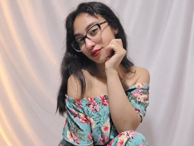 CamiVargas, com cabelo escuro e ondulado, óculos, batom vermelho e um vestido floral com um ombro descoberto, tocando sua bochecha, olhando ligeiramente para o lado, mostrando seus seios de forma sutil..