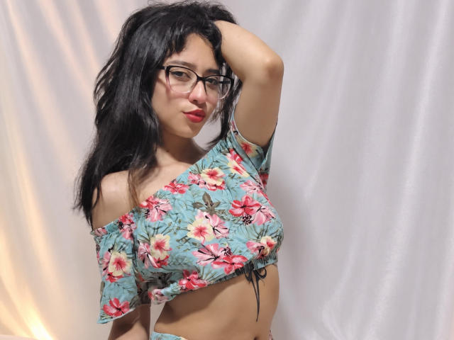 CamiVargas, avec ses cheveux noirs, ses lunettes et son haut à fleurs avec une épaule dénudée, une main dans les cheveux, montrant de petites seins, des lèvres roses et un visage mignon, debout devant un rideau blanc..
