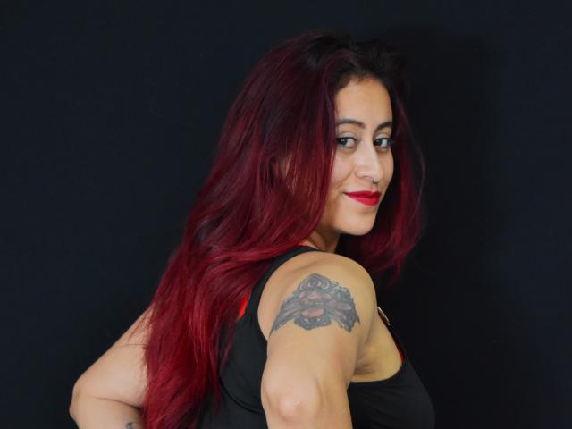 Evans Samantha, com cabelo ruivo e preto, batom vermelho, top preto, tatuagem no braço direito, sorrindo para a câmera, fundo preto, mostrando suas costas e lateral..