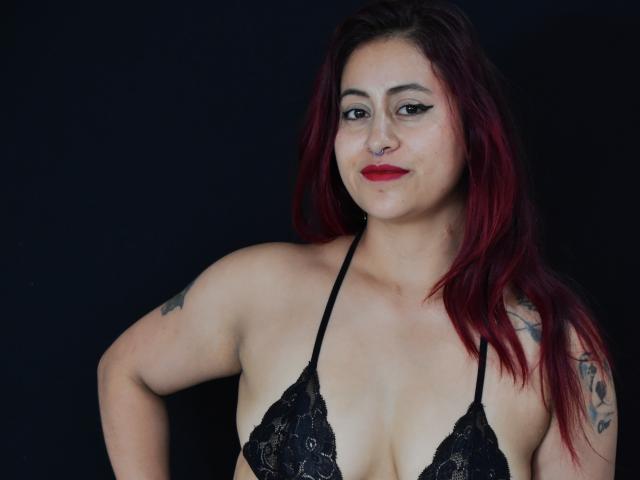 EvansSamantha, met rood haar, een zwarte kanten beha en tatoeages, lachend, waarbij haar borsten zichtbaar zijn, met rode lippenstift, op een donkere achtergrond..