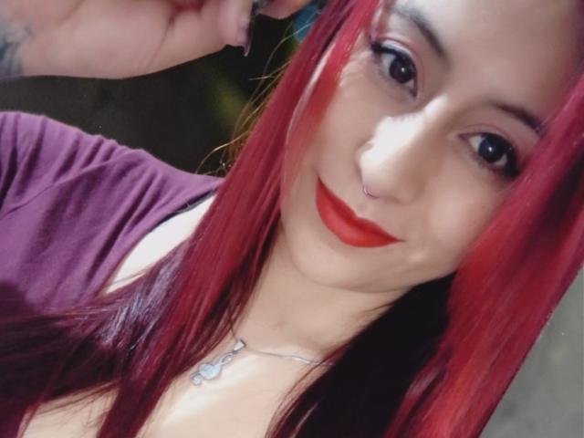 Evans Samantha, con capelli rossi, rossetto rosso, piercing al naso e una collana, indossa una maglietta viola, sorride, mostrando il seno, ha occhi scuri e si tiene i capelli..