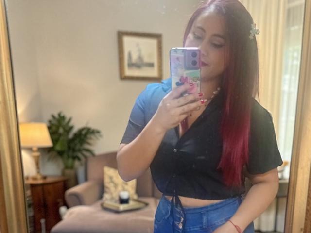 Evans Samantha, com cabelo ruivo, blusa preta e calça jeans, tirando uma selfie no espelho, mostrando seus seios, em um quarto com uma luminária e uma planta..