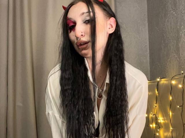 WenMewborn with long black hair, red devil horns, heavy eye makeup, nose ring, and a white shirt, leaning forward with a slight smirk, and string lights in the background.
