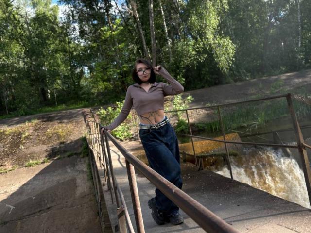 WenMewborn mit Brille, braunem Oberteil und Jeans, steht auf einer rostigen Metallbrücke, die Hand im Haar, und zeigt ihre Tattoos, ihre kleinen Brüste und ihr hübsches Gesicht, bei Tageslicht..