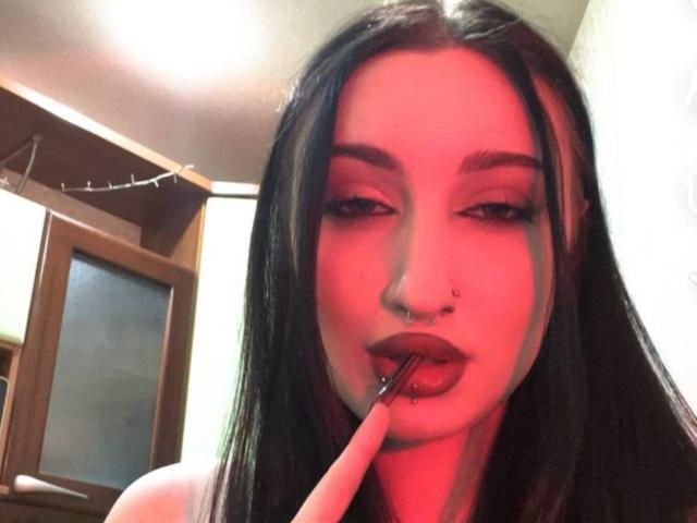 WenMewborn, com cabelos longos e pretos, iluminação vermelha, piercing no nariz e batom escuro, chupando o dedo com a boca ligeiramente aberta, olhando sedutoramente para a câmera..