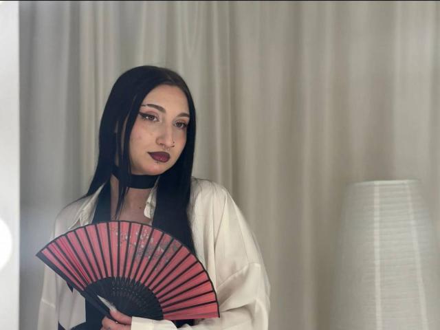 Eine Frau namens WenMewborn mit langem, schwarzem Haar, dunklem Lippenstift und einem weißen Hemd, die einen roten und schwarzen Fächer hält, einen schwarzen Choker trägt und vor einer hohen Lampe steht..