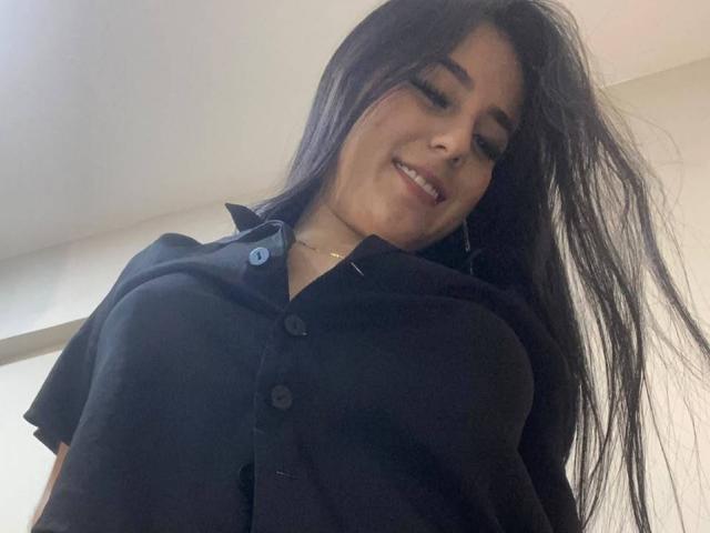 StefanyDelly with long black hair, smiling down, wearing a black button-up shirt, showing her large tits, low-angle shot, slightly messy hair, light skin, subtle makeup.