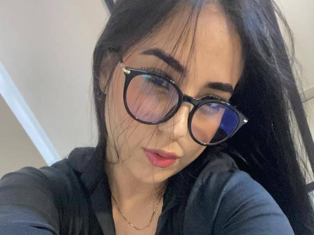 StefanyDelly with long black hair, wearing black glasses and a black shirt, has pink lips and a slight smile, looking into the camera with a subtle, sexy expression.