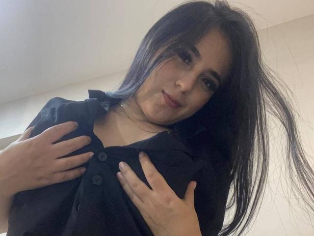 StefanyDelly with long black hair, wearing a black shirt, hands on her tits, slightly smiling, looking down at the camera, light skin, subtle makeup, indoor setting.