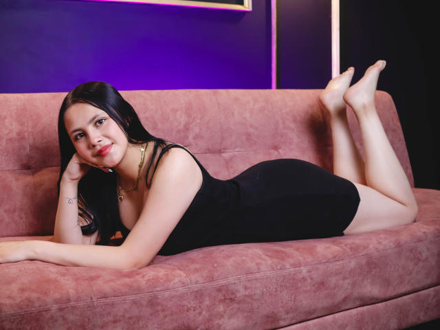 AllyJhones, sdraiata su un divano rosa in pelle scamosciata, indossa un abito nero aderente che mette in mostra il suo sedere, ha lunghi capelli neri, sorride, ha le gambe sollevate, piedi nudi e indossa una collana d'oro..