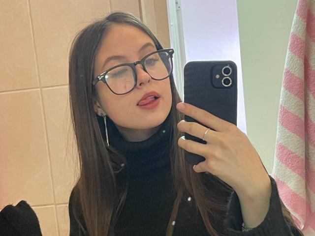 Audrie Nuessen mit Brille, die Zunge leicht herausgestreckt, trägt einen schwarzen Rollkragenpullover, hält ein schwarzes Telefon, zeigt einen Ring an ihrer linken Hand und hat glattes, braunes Haar..