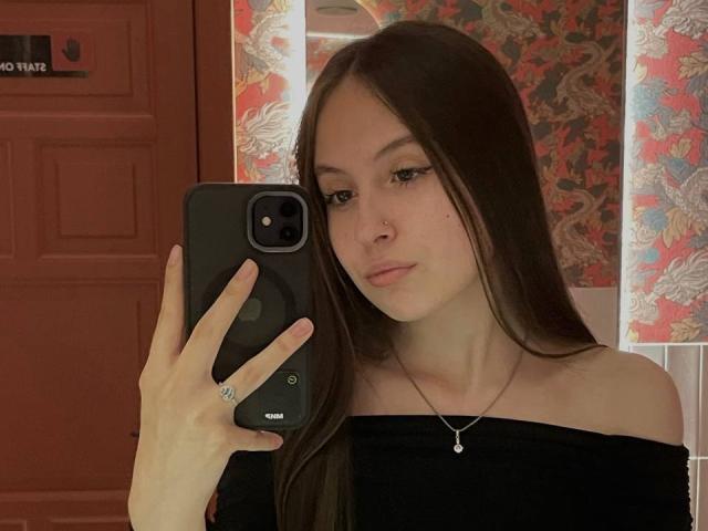 Audrie Nuessen, con lunghi capelli castani, indossa una maglietta nera con una spalla scoperta, tiene in mano un telefono nero, si sta guardando, con un trucco leggero, ha un naso piccolo, indossa una collana e un anello, sullo sfondo si vede una porta rossa..