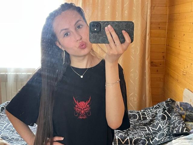 Audrie Nuessen met lang bruin haar, een opgetrokken mond, een zwart shirt met een rood ontwerp, die een telefoon vasthoudt, staand in een slaapkamer, met een houten muur, een bed met een patroon, een zilveren ketting, een armband, een schattig gezicht..