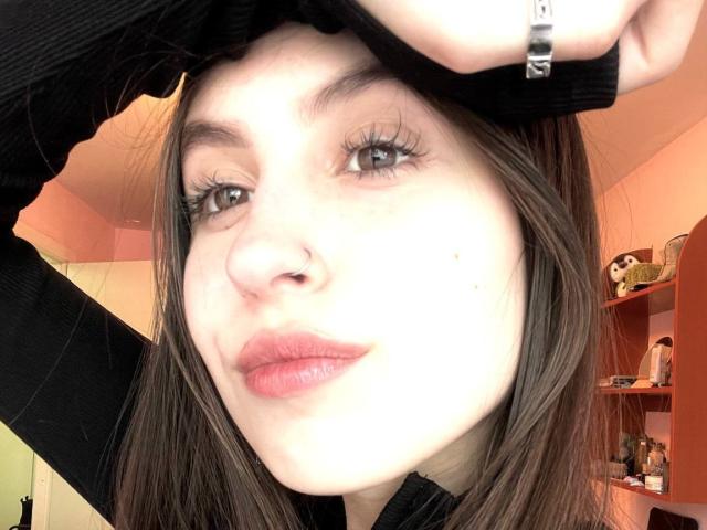 Audrie Nuessen, com seus lábios rosados e curvados, olhos castanhos e piercing no nariz, levantando uma camisa preta, exibindo seus longos cílios e uma pequena pinta na bochecha esquerda..