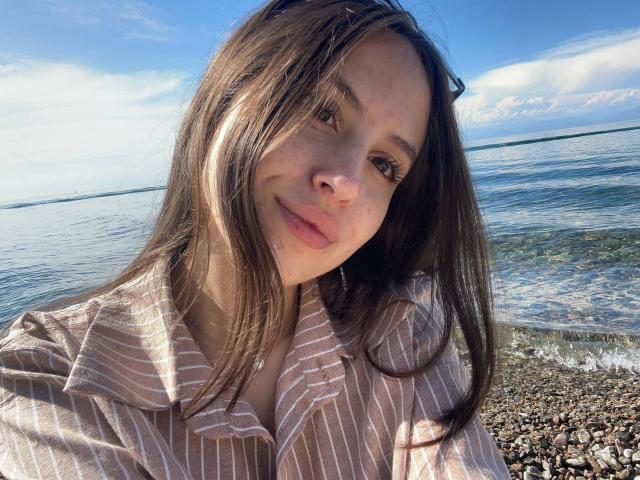 Audrie Nuessen, con cabello castaño, vestida con una camisa a rayas, tiene senos pequeños, una cara linda y una sonrisa sutil, y está de pie junto a una playa de guijarros con un cielo azul claro..