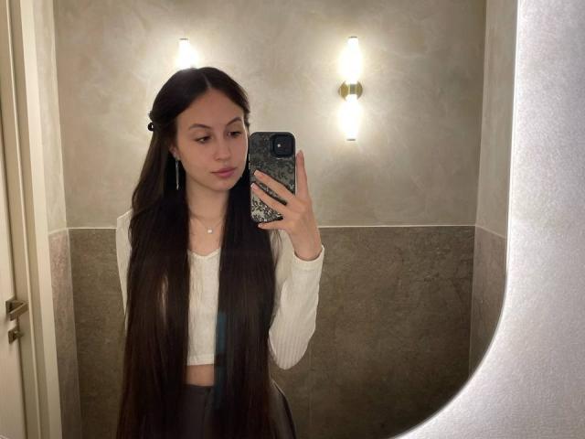 Audrie Nuessen, con lunghi capelli castani, indossa una canottiera bianca e una gonna nera, e sta scattando un selfie allo specchio in un bagno, tenendo in mano un telefono con una custodia scura a motivi..