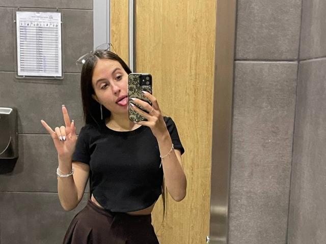 Audrie Nuessen, avec de longs cheveux bruns, un crop top noir laissant entrevoir de petites seins, une jupe noire, la langue tirée et faisant un signe de la paix, tenant un téléphone pour un selfie devant le miroir dans une salle de bain..