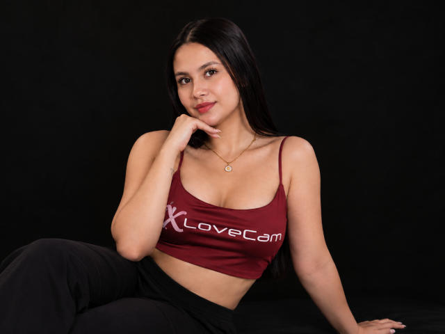 AllyJhones mit langem, schwarzem Haar, braunen Augen und heller Haut, trägt ein rotes LoveCam-Top und schwarze Hosen, sitzt vor einem schwarzen Hintergrund und berührt ihr Kinn..