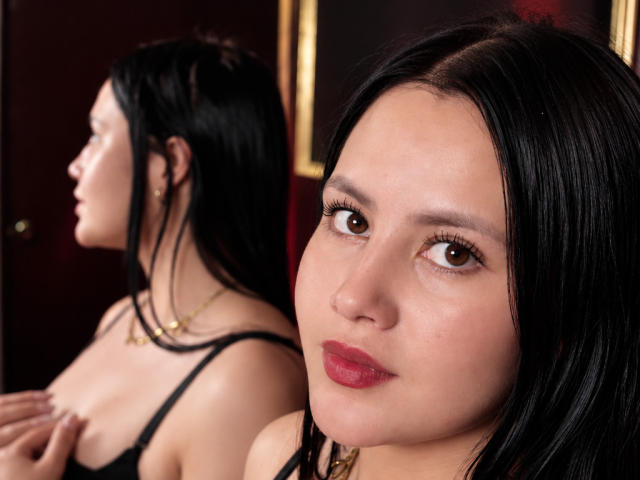 AllyJhones, met zwart haar en rode lippenstift, bruine ogen en een lichte glimlach, staat voor een spiegel die haar blote rug en zwarte beha weerspiegelt, waarbij haar borsten zichtbaar zijn..